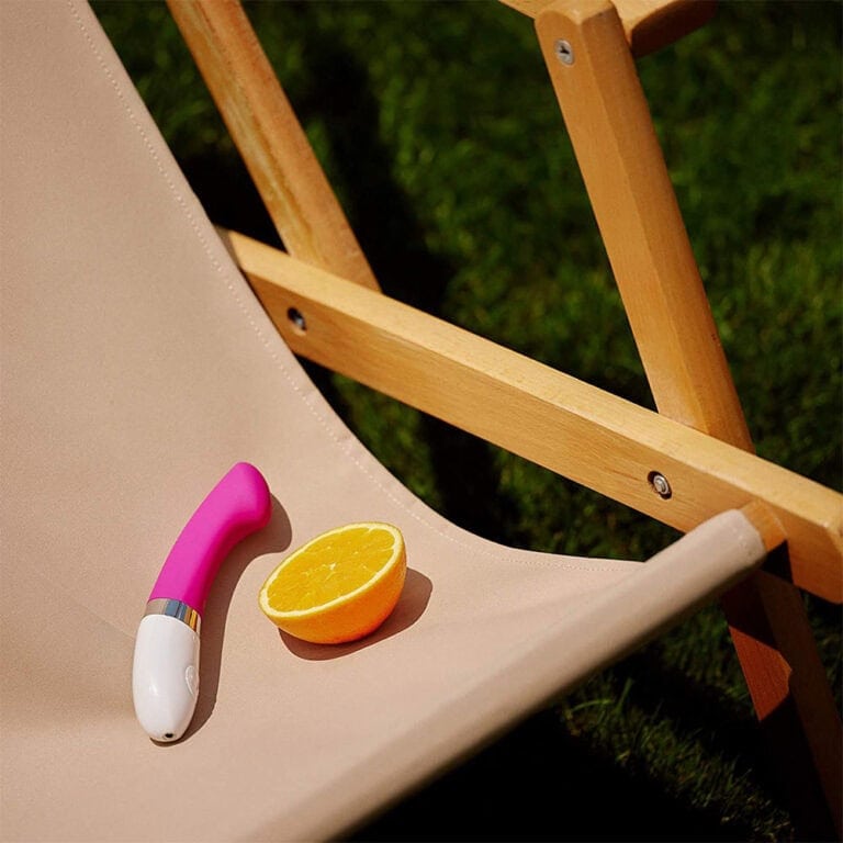 Sun lounger with a pink vibrator and a fresh orange slice on a beige fabric surface, outdoors on a sunny day. Perfect for exploring pleasure and relaxation with high-quality sextech products.